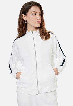 Jonge vrouw met bruin haar draagt een witte jas met rits en zwarte strepen op de mouwen, handen in de zakken, kijkt naar links.
