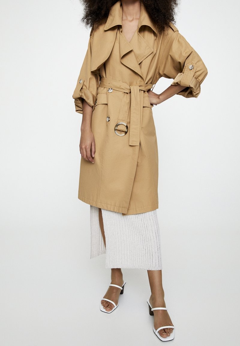 Tan trenchcoat met een riem en zilveren hardware, met wijde mouwen en een getailleerde pasvorm. Draagt over een witte geribde rok en sandalen met bandjes.