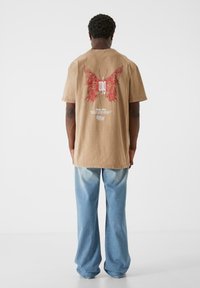 Brown cotton t-shirt with red butterfly graphic on the back, relaxed fit, and short sleeves; paired with light blue wide-leg jeans.