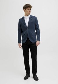 Tumesinistega bleiser tekstureeritud kangast, ühe nupuga kinnitamine, kahe taskuga ja kontrastsete servadega, koos mustade pükste ja mustade kingadega.