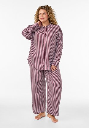 Mujer con cabello rizado, vestida con un conjunto de pijama holgado a rayas a juego con rayas verticales rojas y blancas, de pie descalza, con la mano en el cuello, sonriendo.