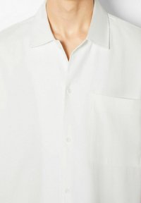 Camicia bianca a maniche corte con bottoni realizzata in tessuto morbido. Presenta un colletto a punta e una tasca sul lato sinistro del petto. Texture satinata sottile.