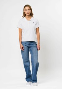 Hvitt kortermet pikéskjorte med teksturert, ribbet design og en liten logo på venstre bryst, kombinert med løse blåjeans og hvite joggesko.