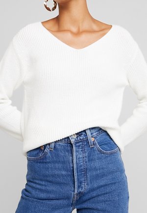 Frau trägt einen weißen V-Ausschnitt-Pullover aus Strick, der in hoch taillierte blaue Jeans gesteckt ist, mit großen sichtbaren Creolen.