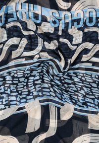 Lehký šátek z tkaniny s abstraktními modrými a béžovými vzory na tmavém pozadí. Vlnité linky a tečkované vzory vytvářejí texturu a vizuální zajímavost.