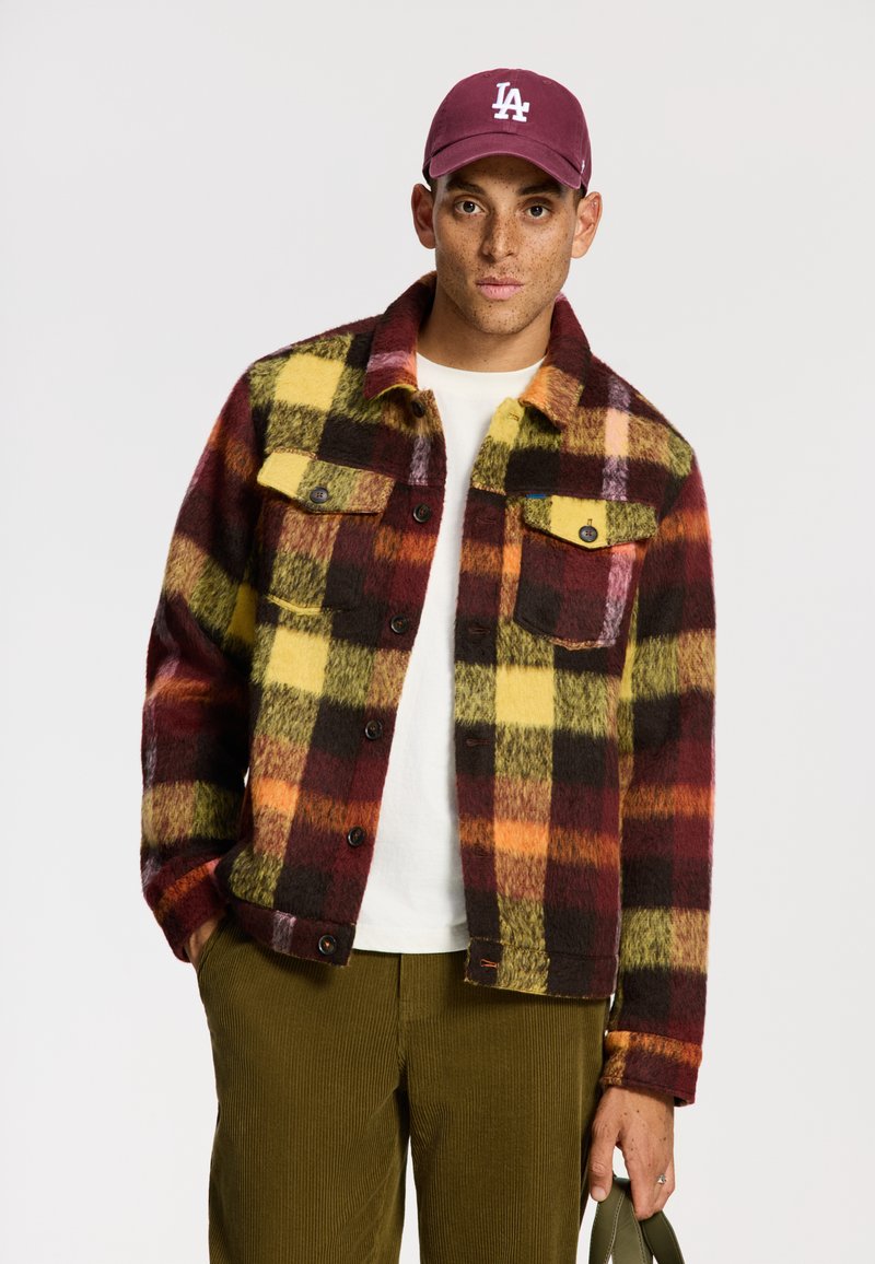 Giacca in pile a righe gialle, bordeaux e verdi, con due tasche sul petto e bottoni neri, indossata sopra una camicia bianca e pantaloni oliva.