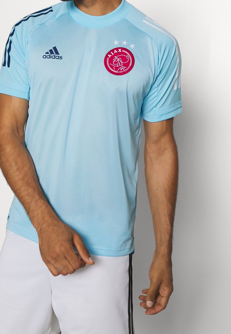 Jersey atlético de color azul claro con mangas cortas, logo de Adidas, emblema del Ajax y tres rayas blancas en cada hombro. Tejido texturizado.