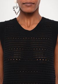 Czarny, dzianinowy top bez rękawów z teksturowanym, otwartym splotem. Posiada żebrowany dekolt i subtelny połysk. Widoczne srebrne kolczyki w kształcie serca.