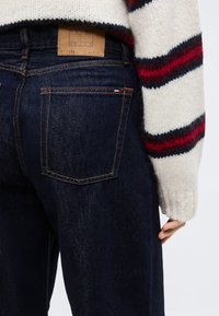 Σκούρο μπλε τζιν παντελόνι με πίσω τσέπη και μικρό λογότυπο σημαίας, συνδυασμένο με λευκό πλεκτό πουλόβερ που έχει κόκκινες και ναυτικές ρίγες στο μανίκι.