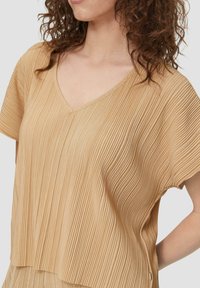 Beige plissé top met V-hals, korte mouwen en textiel met structuur. Heeft een losse pasvorm en een zijsplit voor extra detail.