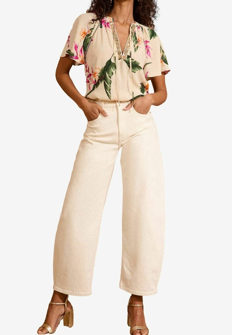 Beige Bluse mit Blumenmuster, kurzen Ärmeln, V-Ausschnitt und Paillettenverzierungen, kombiniert mit hoch taillierten, weit geschnittenen cremefarbenen Jeans und goldenen Riemchen-Heels.