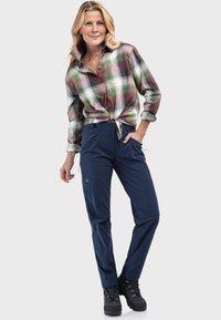 Karierte Hemd in Grün, Lila und Weiß, in der Taille gebunden; navyblaue Hose mit Taschen; schwarze, geschnürte Wanderstiefel.