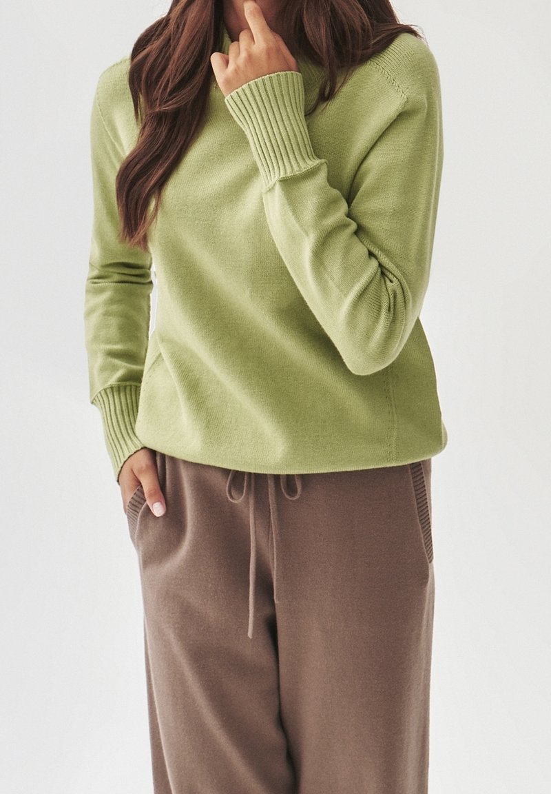 Hellgrüner Pullover mit gerippten Bündchen, Rundhalsausschnitt und weicher Textur. Kombiniert mit braunen Jogginghosen mit Kordelzug und seitlichen Taschen.