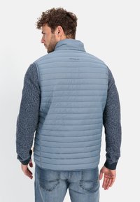 Light blue puffer vest with horizontal stitching, high collar, and textured dark blue sleeves. Features tag on collar and casual fit.