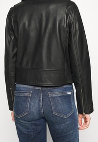 Černá kožená krátká bunda s přiléhavým střihem, výrazným štepováním a zipy na rukávech, nošená přes modré džínové kalhoty.