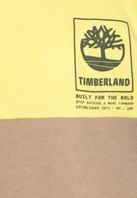 T-shirt in cotone giallo e marrone con un grafico del logo Timberland, caratterizzato da testo in grassetto e un design di albero. Maniche corte, collo rotondo.