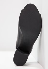 Zwart leren schoen met een spitse neus en een gladde oppervlakte, voorzien van een getextureerde rubberen zool voor grip, schuin gepositioneerd tegen een witte achtergrond.