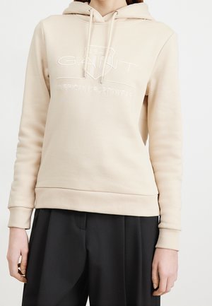 Beżowa bluza z kapturem z troczkami i logo "GANT American Sportswear" noszona z czarnymi spodniami na osobie, pokazująca górną część tułowia i ramiona.