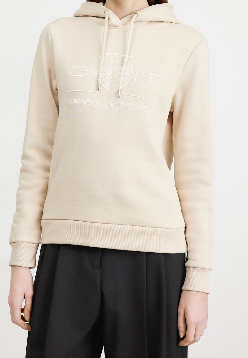 Sweat à capuche beige avec cordons et logo "GANT American Sportswear" porté avec un pantalon noir sur une personne, montrant le haut du torse et les bras.