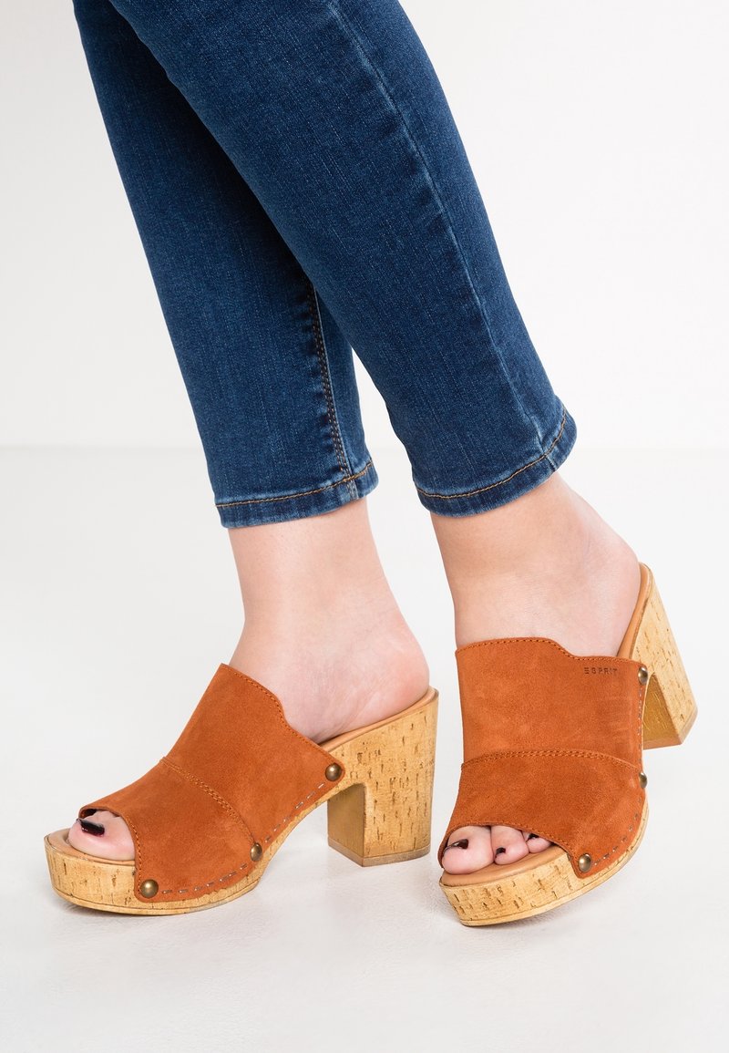 Mules ouvertes en daim marron avec talons compensés en liège épais, présentant un design supérieur courbé et des détails de surpiqûres contrastantes.