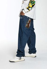 Baggy blue denim jeans with unique patterned fabric on the back pocket; paired with white sneakers featuring black and gray accents.