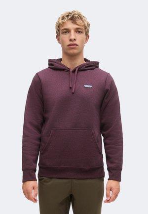 Joven con cabello rubio corto lleva una sudadera con capucha de color morado oscuro con bolsillo delantero y pantalones marrones, de pie contra un fondo claro.