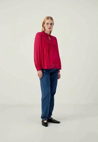 Blusa vermelha de manga longa com decote franzido, tecido texturizado e pregas. Combinada com jeans de denim azul e sapatos pretos.
