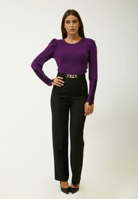Lila gerippter Langarm-Top mit gepolsterten Schultern; hochtaillierte schwarze Hose mit vertikalen Nähten; goldene Ketten-Gürtelschnalle; schwarze spitze Absatzschuhe.