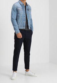 Mann in einer hellblauen Jeansjacke, dunkler Hose, dunklem Hemd und weißen Sneakers, der vor einem schlichten hellen Hintergrund steht.