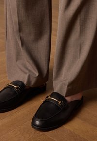 Black leather slip-on shoes with a gold metal accent, paired with light brown, loose-fitting, textured trousers. Wood flooring visible.