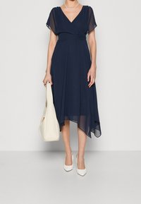 Mörkblå klänning med korta, genomskinliga ärmar och en omslagdesign. Har en asymetrisk fåll och matchas med vita pumps med spetsig tå och en ljus handväska.