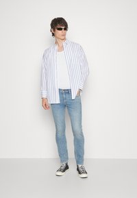 Camisa blanca de botones con rayas verticales azules, superpuesta sobre una camiseta sin mangas blanca. Jeans ajustados de color azul claro y zapatillas altas negras.