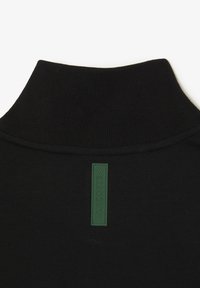 Sort sweater med en tekstureret strikkrave. Har en rektangulær grøn gummimærke syet på bagsiden, som tilføjer detaljer til designet.
