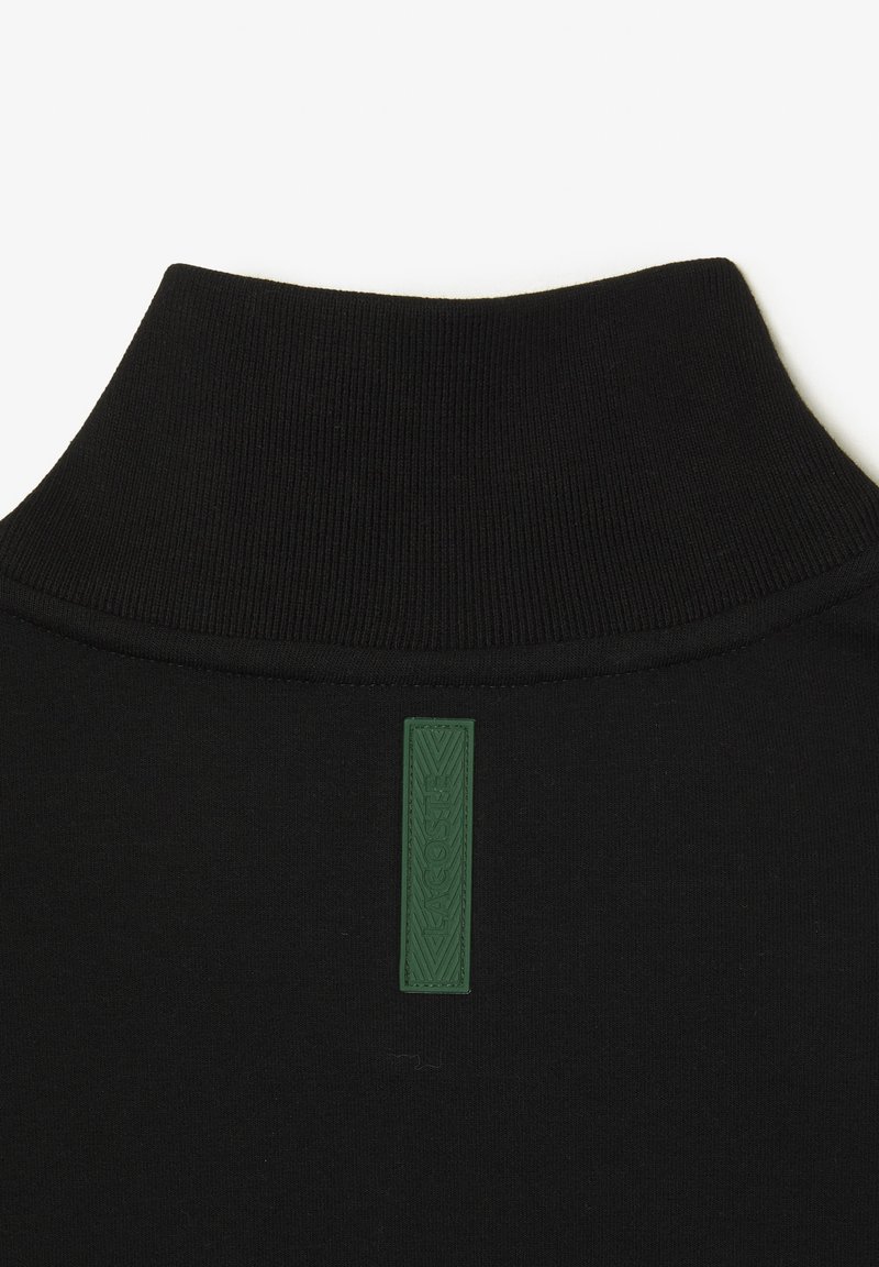 Maglione a collo alto nero con colletto a maglia testurizzata. Presenta un'etichetta rettangolare verde in gomma cucita sul retro, che aggiunge un tocco di stile al design.