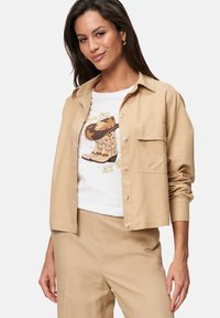 Lichte button-up overhemd met een borstzak, gedragen over een wit T-shirt met een grafisch ontwerp van een laars, gecombineerd met bijpassende lichte broek.