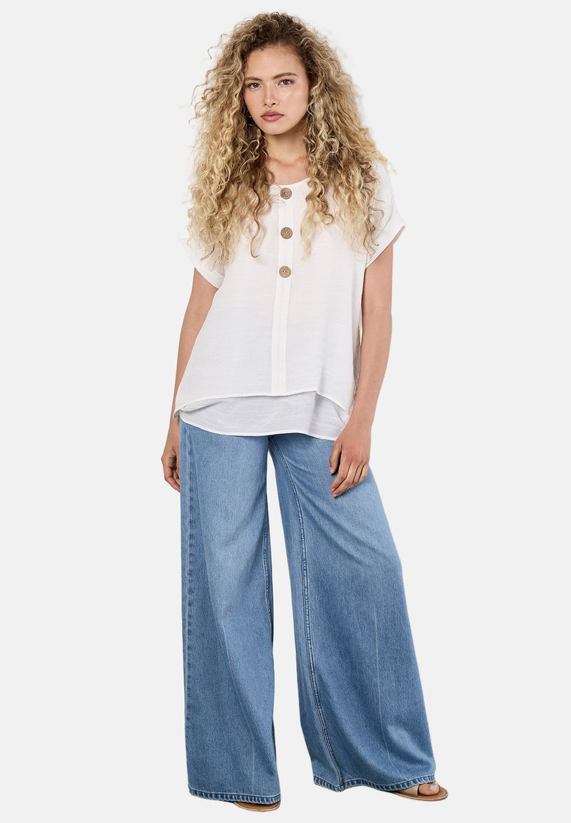 Femme aux cheveux bouclés blonds portant une blouse blanche à manches courtes boutonnée et un jean large bleu clair, debout devant un fond uni.