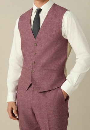 Homme portant un gilet et un pantalon de costume grenat, une chemise blanche et une cravate noire, avec une main dans la poche.