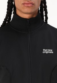Svart glidelåsjakke med høyt krage laget av glatt stoff, med hvit brodert tekst "the new originals" på venstre brystområde.
