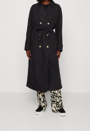 Person wearing a long black trench coat with belt, patterned black and white wide-leg pants, and black slip-on shoes with white soles.