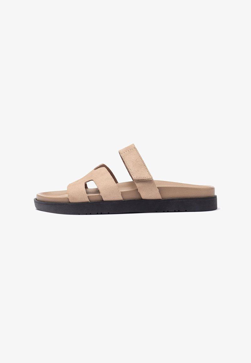 Beige suede slide sandals with a black rubber sole, featuring two wide straps and a cut-out design for ventilation and comfort.