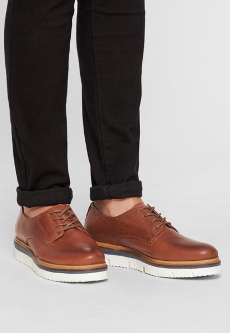 Personne portant des chaussures en cuir marron à lacets avec des semelles blanches épaisses et un pantalon noir retroussé sur un sol gris clair.