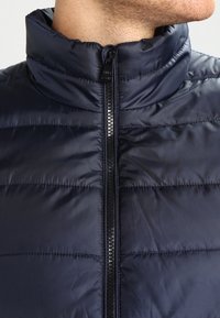 Laivastonsininen toppaliivi, jossa on korkeakaulus, etuvetoketju ja vaakasuora tikattu kuvio. Sileä, kevyt kangas kiiltävällä viimeistelyllä.