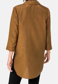 En kvinna ses bakifrån, iförd en brun, silkeslen, långärmad skjorta och svarta byxor, med handen vilande på höften.