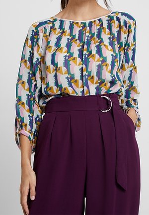 Femme portant un chemisier coloré à motifs abstraits, rentré dans un pantalon violet plissé taille haute avec une ceinture à boucle argentée.