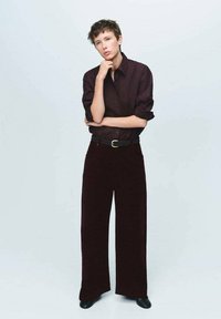 Camisa burdeos con mangas enrolladas, combinada con pantalones anchos burdeos de terciopelo. Un cinturón negro y zapatos completan el look.