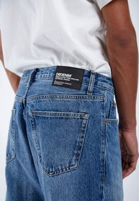 Light blue denim jeans featuring a woven texture, standard pockets, and a black label with white text on the back waistband.