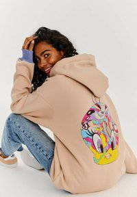 Béžová mikina s farebnou kreslenou grafikou na chrbte, dlhé rukávy s levanduľovými akcentmi, v kombinácii so svetlomodrými džínsami a bielymi teniskami.