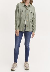 Kvinna som bär en ljusgrön knapplåst jacka med dragkedjefickor, blå skinny jeans och beige sneakers, stående mot en vit bakgrund.