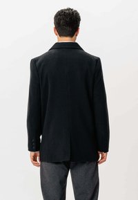 Mand set fra ryggen, iført en sort blazer med knaplukning på ærmerne og mørkegrå bukser mod en ensfarvet lys baggrund.