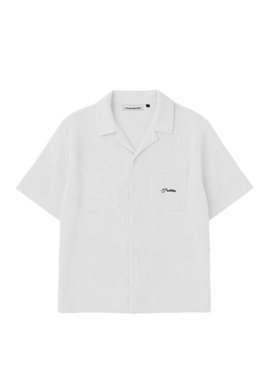 Femme portant une chemise blanche texturée à manches courtes boutonnée sur un haut et un pantalon assortis, avec une casquette de baseball bleu marine, les mains dans les poches, regardant vers le bas.